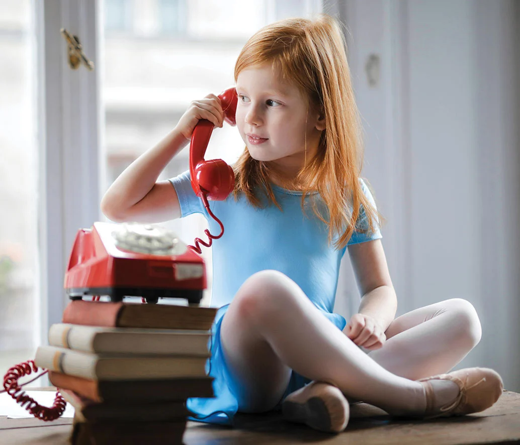 A young red haired girl with her legs crossed sits on a table talking on a red dial-up phone that is on top of a stack of books.