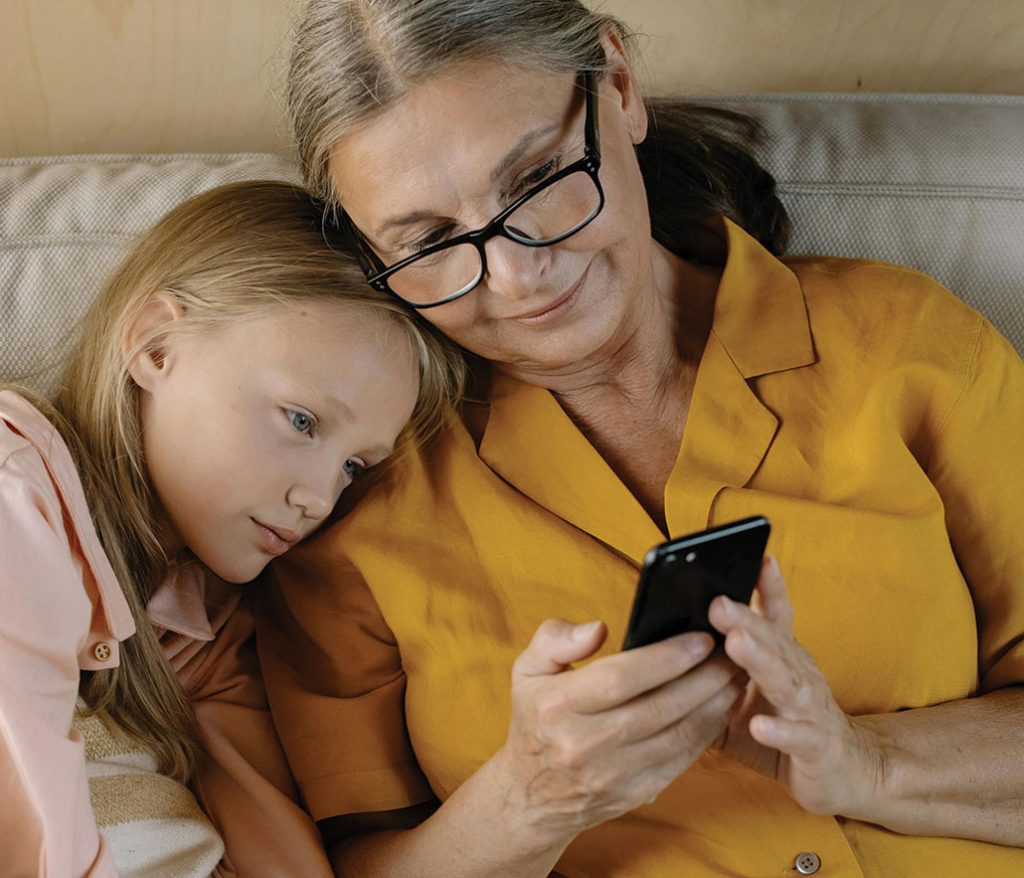 Grandmother shows her granddaughter something on her phone while smiling and cuddling on the couch.