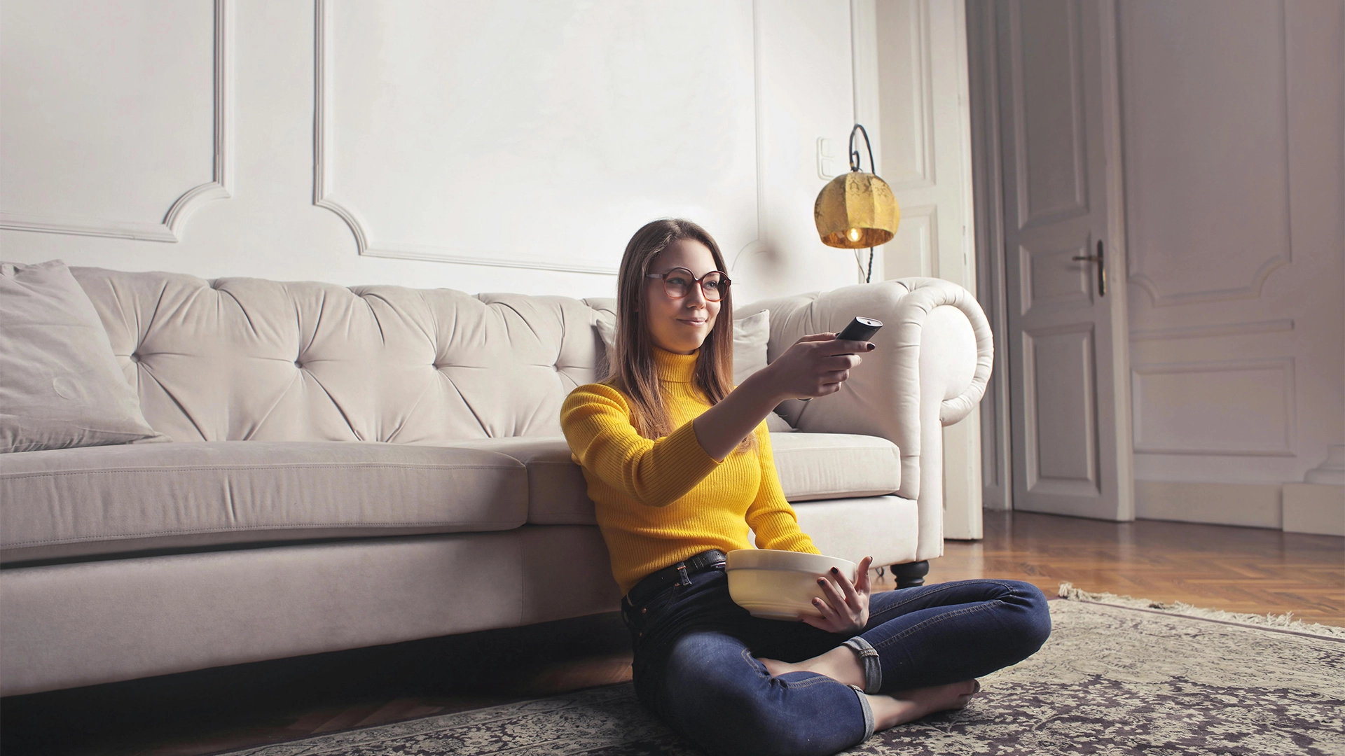 Woman sitting on the floor against a white sofa pointing a TV Remote at presumably her Television