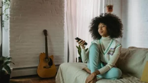 Woman sitting cross legged on a grey sofa holding a remote to presumably a television